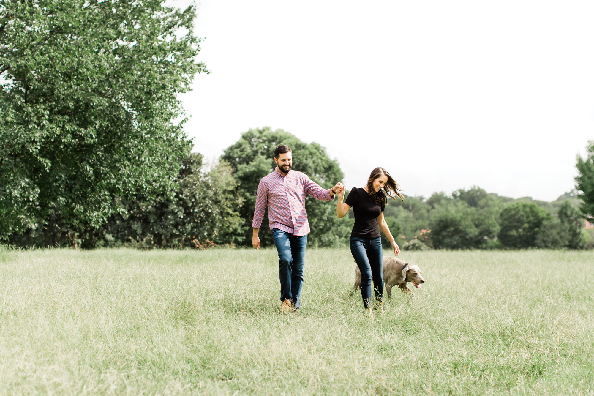 Engaged couple walks their dog through a grassy field